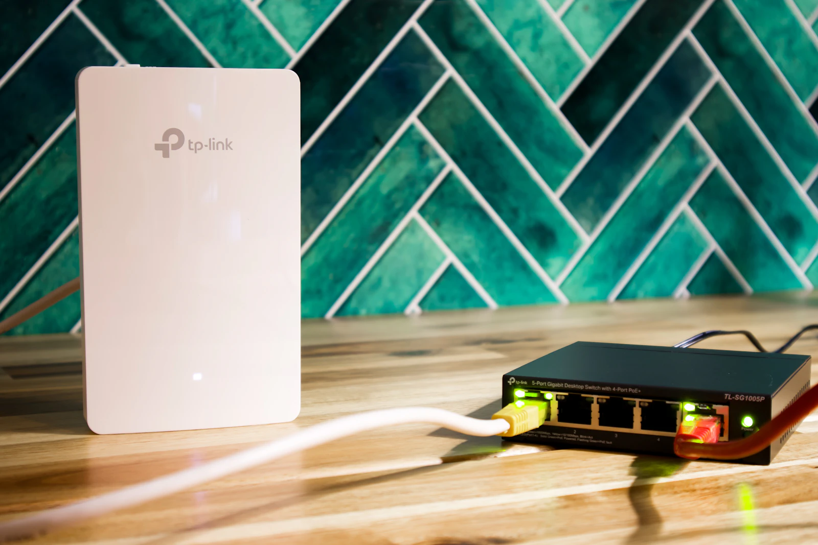 White Wi-Fi access point beside a small black network switch with lit Ethernet ports and yellow and red cables on a wooden counter