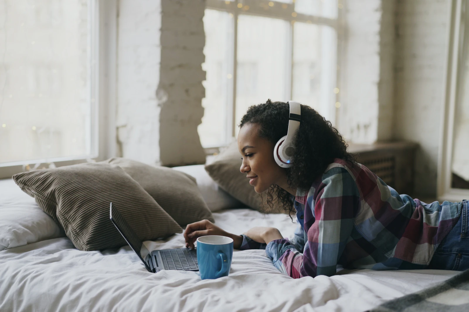 Young woman wearing headphones lying on a bed by a bright window, watching a laptop screen with a blue mug beside her