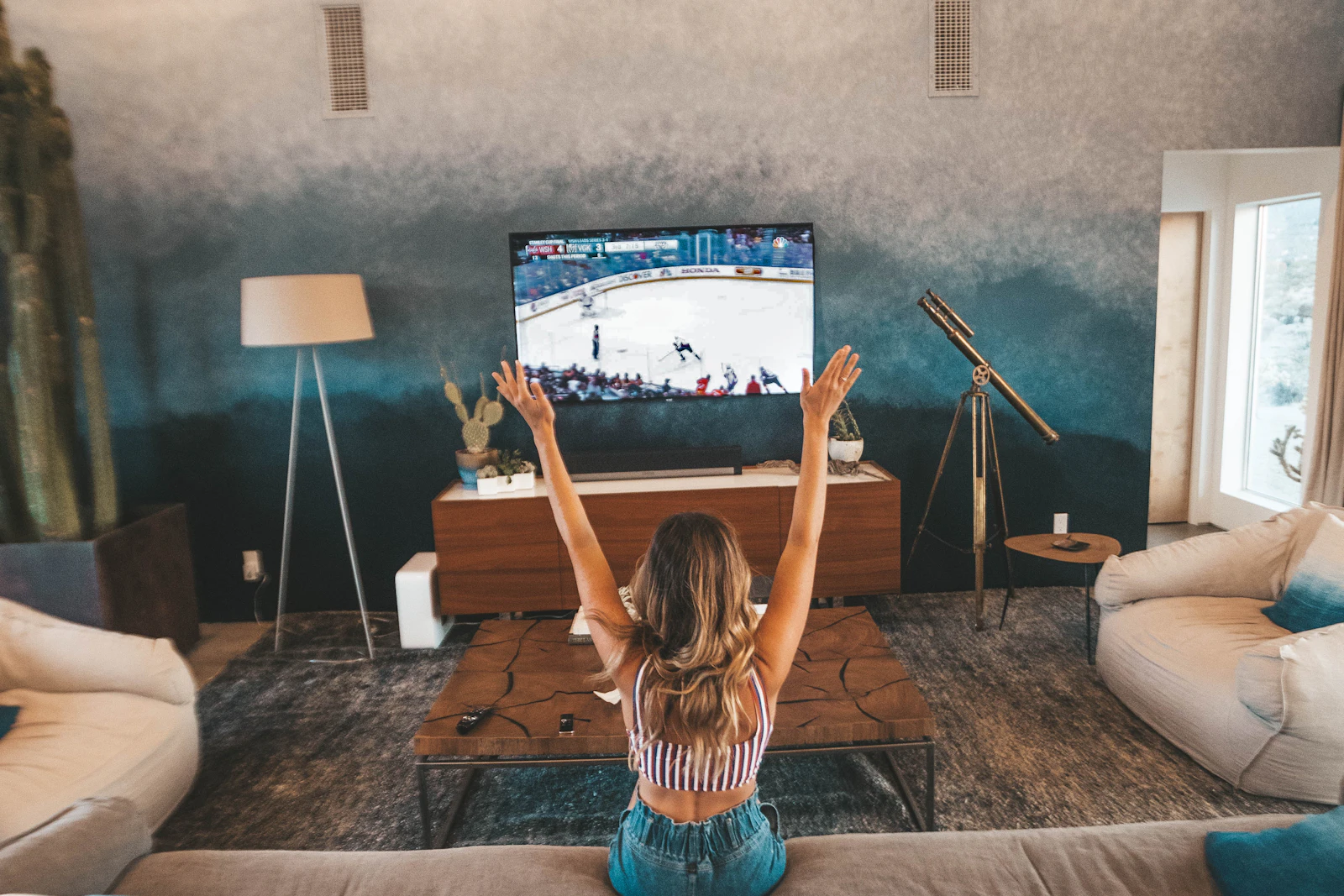 Woman seen from behind on a sofa raising both arms toward a wall-mounted TV showing a live hockey broadcast in a spacious living room